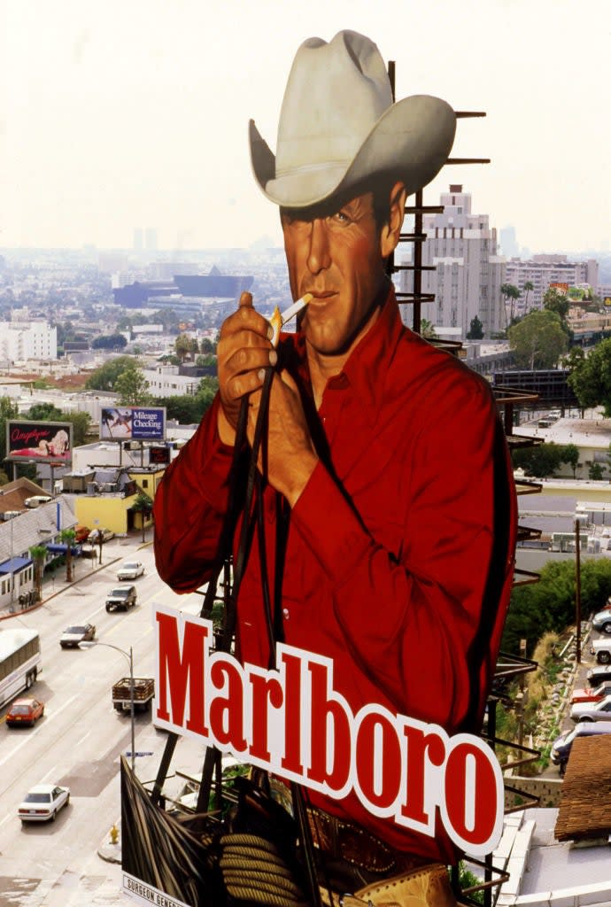 Marlboro billboard with a cowboy, wearing a cowboy hat and collared shirt, holding a cigarette, overlooking a city with buildings and cars