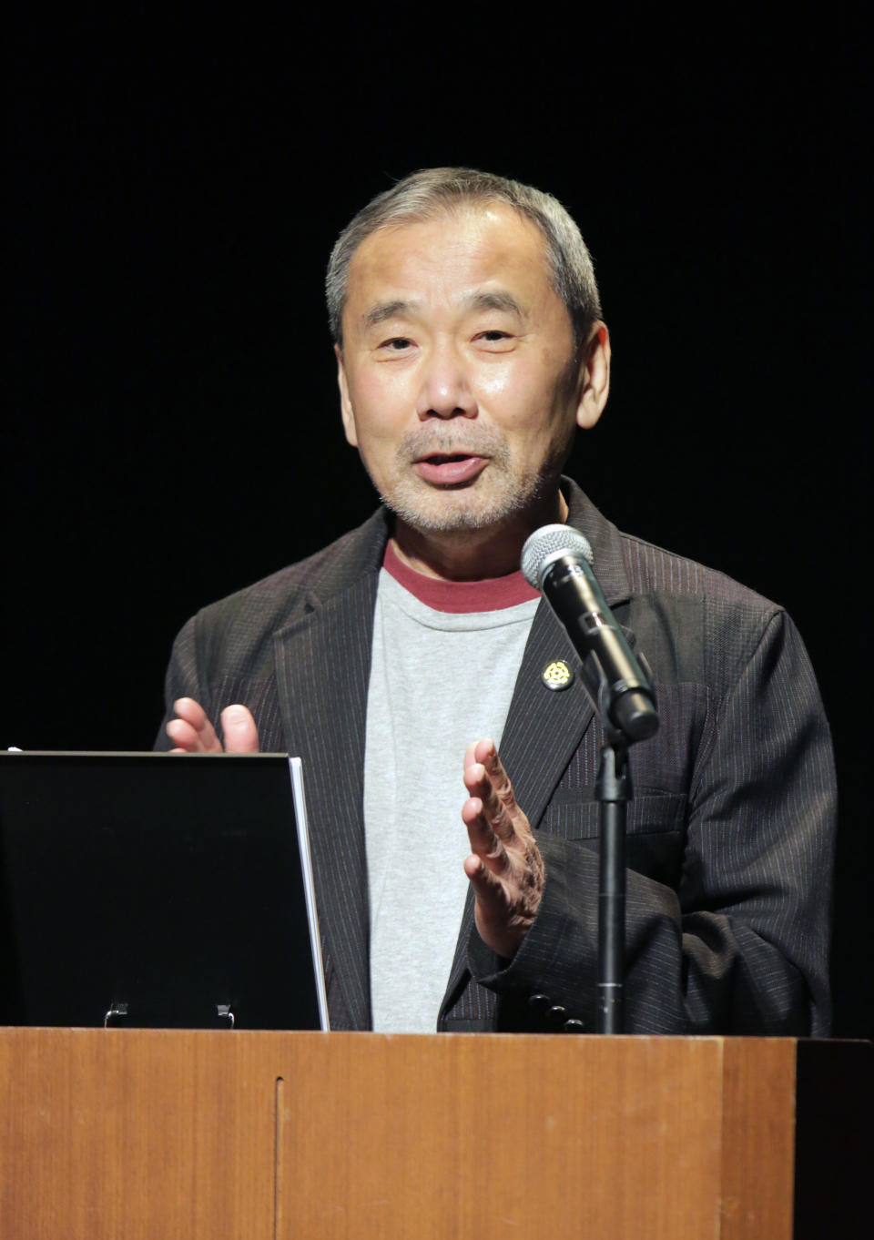 In this photo provided by Shinchosa, author Haruki Murakami speaks on stage in Tokyo, Tuesday, Dec. 17, 2019. A monkey that confesses he steals women's identity cards, causing them to temporarily forget who they are, stars as author Haruki Murakami marks 40 years since his debut as a novelist with his first public reading in Japan in nearly a quarter century. Now 70 and one of the world’s most popular and acclaimed novelists, Murakami debuted with “Hear the Wind Sing” in 1979, four years after he began writing while running a jazz bar in Tokyo. (Shinchosa via AP)