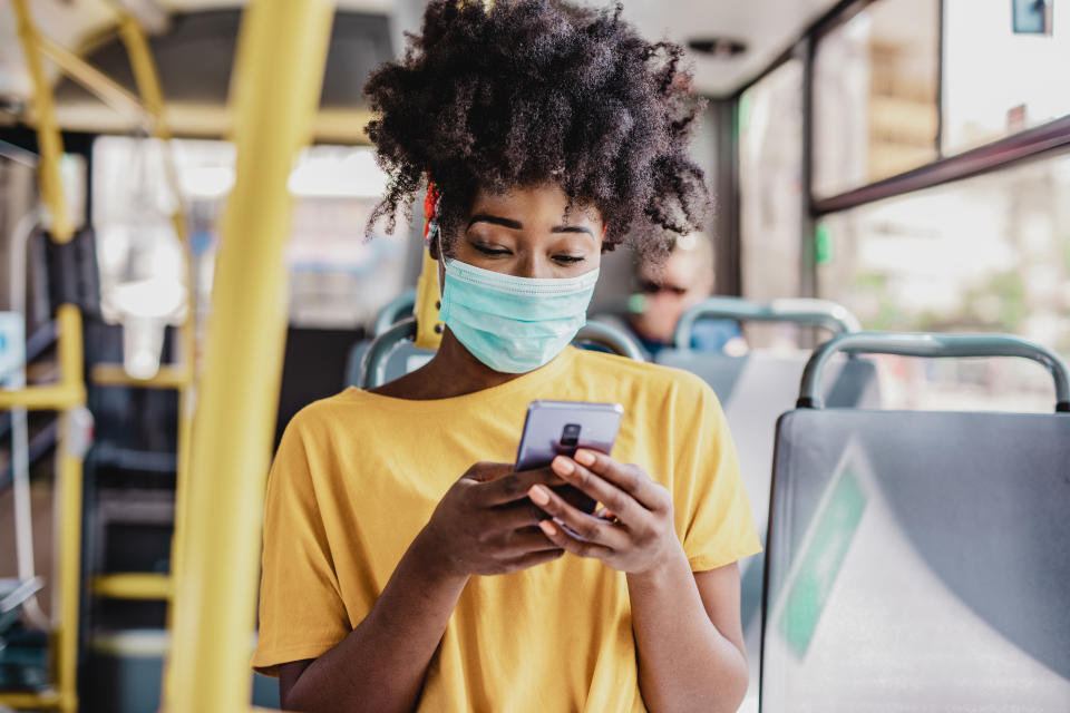 African American woman wearing a protective mask while sitting in a bus and using a mobile phone