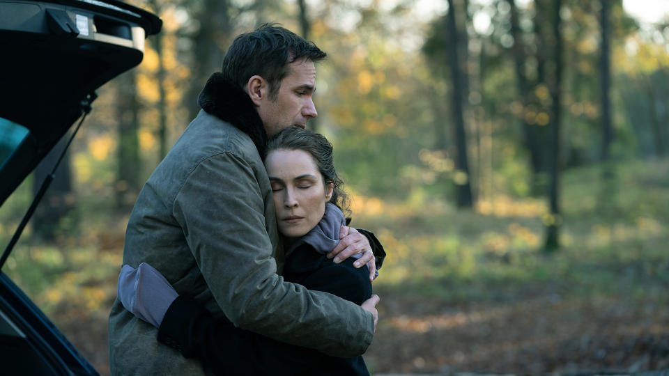 Husband and wife hugging. The couple are wearing warm clothing. In the background you can see woods and an open back door of a car.