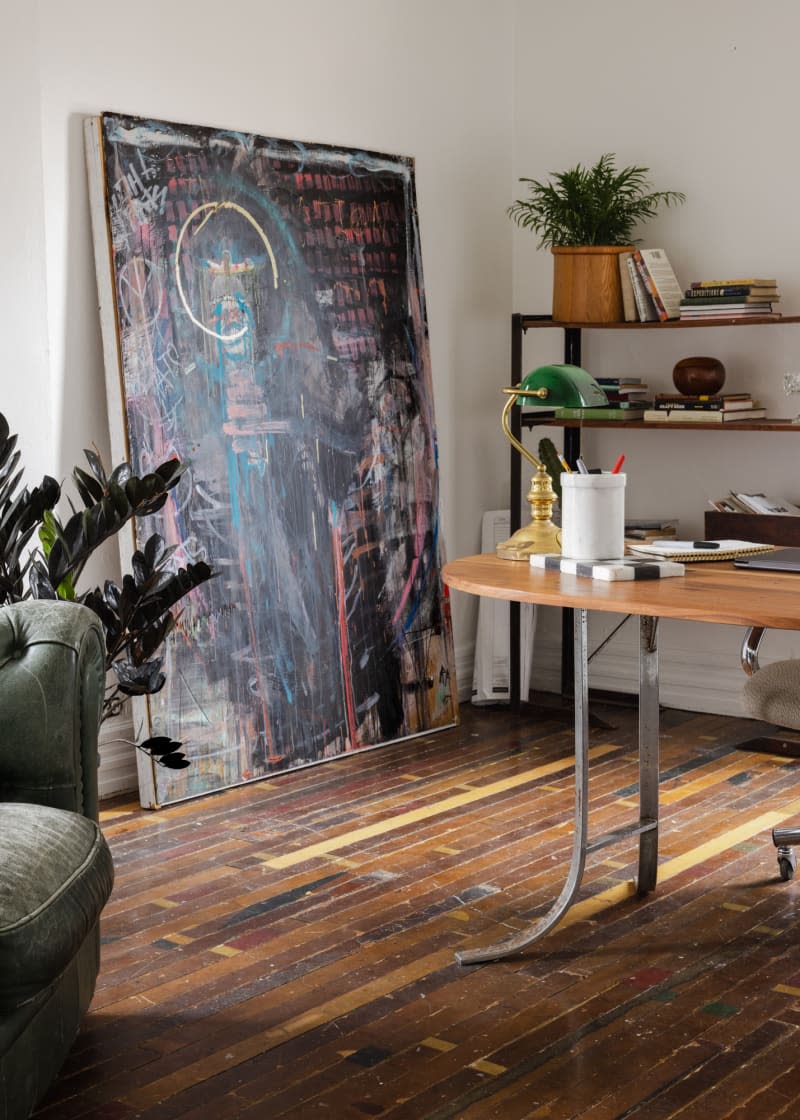 Large artwork leaning against wall of room with plant on shelving unit and wood flooring.