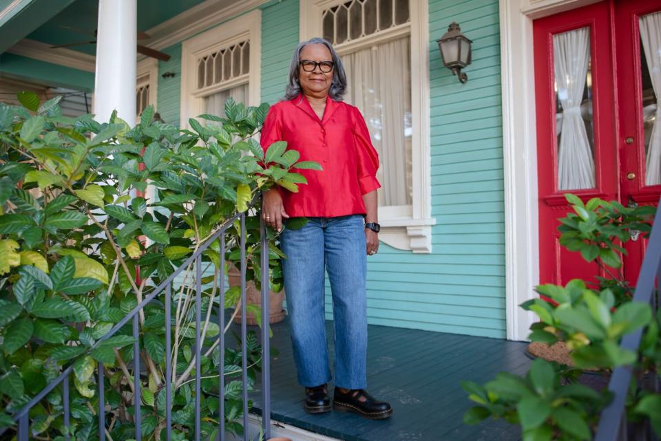 A woman in a red blouse and blue jeans poses on the porch of a house.