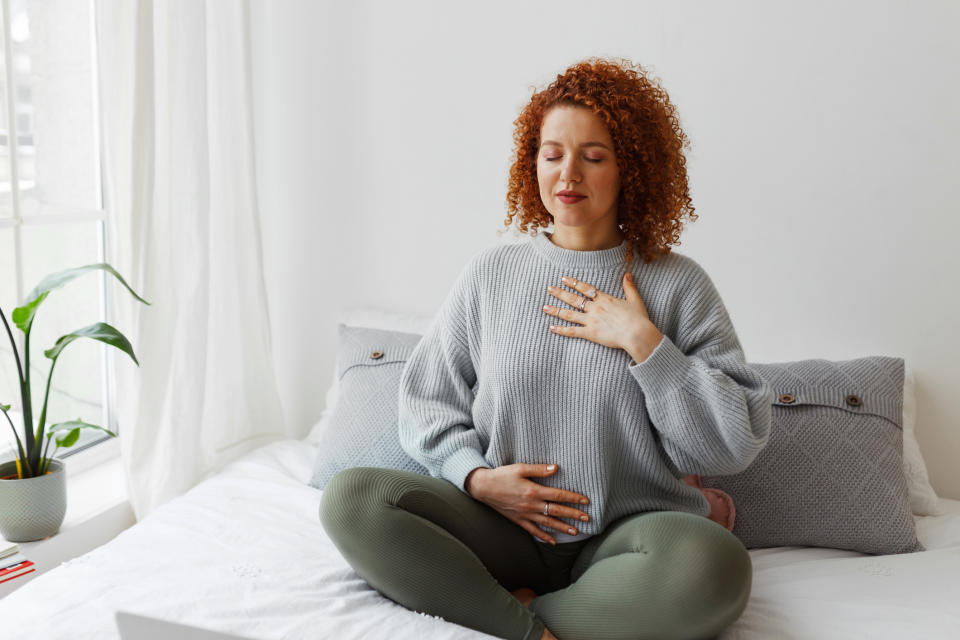 A person with curly red hair sits cross-legged on a bed, eyes closed, with one hand on their chest and the other on their abdomen, appearing to meditate