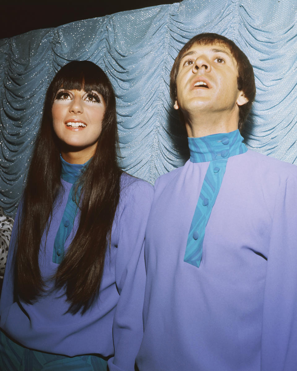 Two people wearing matching blue turtlenecks with scarves against a textured background