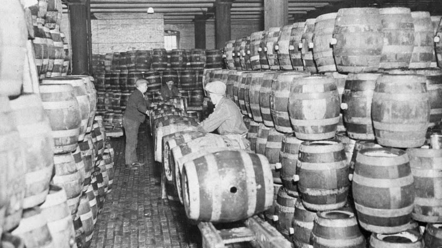 Two men pound spigots into barrels of beer in a large warehouse lined with barrels.