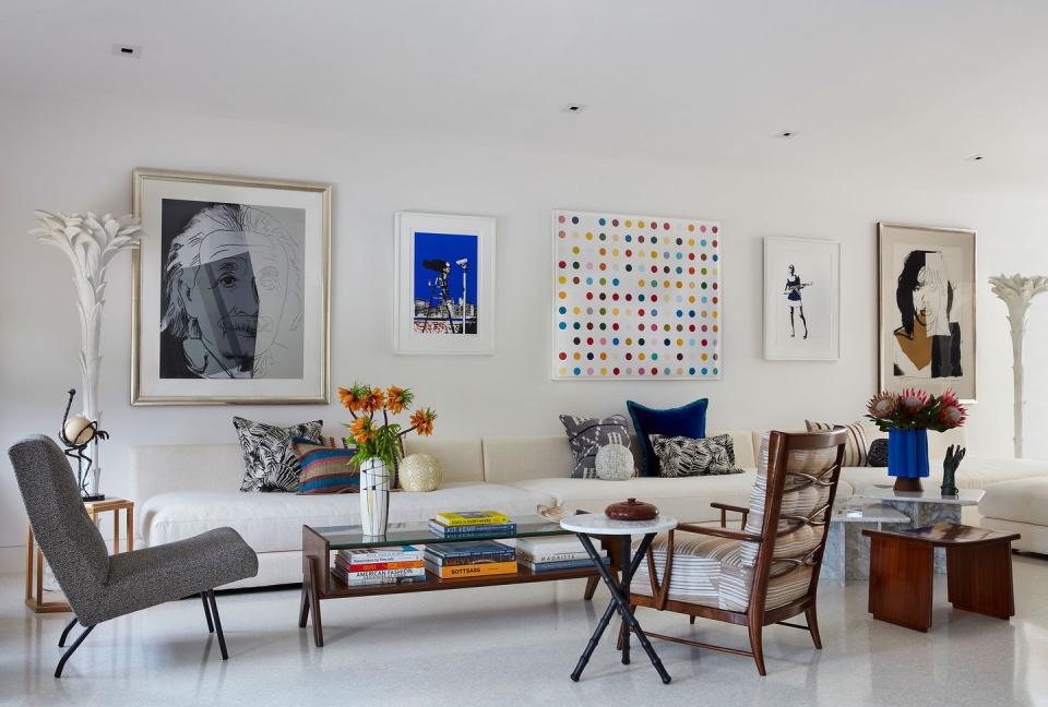 living room with glossy white interior, long white cushioned sofa along the wall and several chairs and low tables in front and five pieces of art hanging on the wall above the sofa, one of which is a large modernistic sketch of albert einstein on the far left