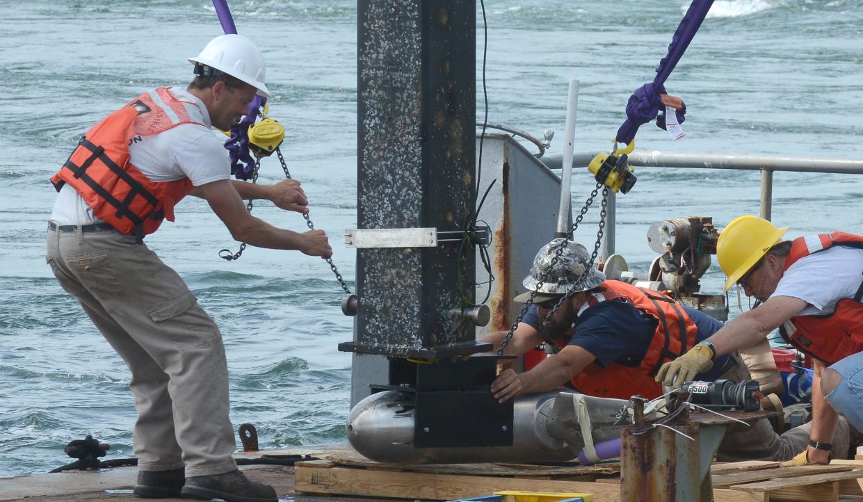 A torpedo-looking tidal turbine is bolted into place in 2021 on a lift arm on a platform just west of the railroad bridge on the Cape Cod Canal in Buzzards Bay. The turbine and several attached sensors were being installed as part of ongoing research into tidal power.