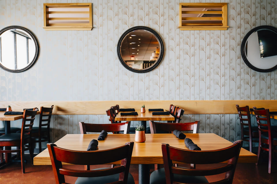 An empty restaurant with wooden tables and chairs, black napkins on each table, decorative round mirrors, and a small potted plant in the center of the image