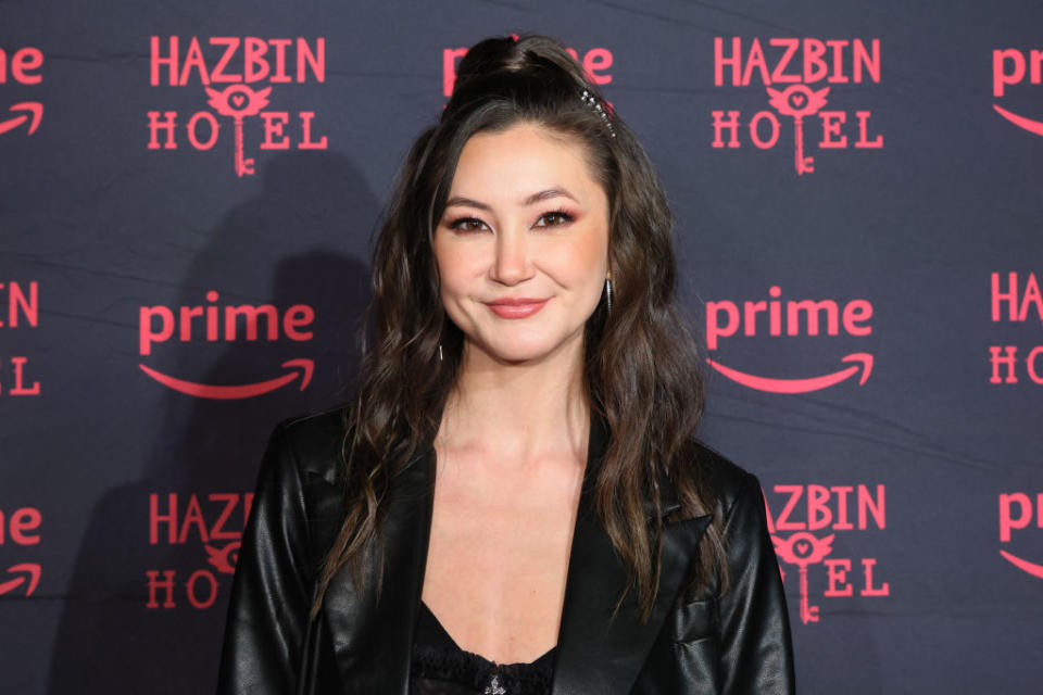 Kimiko Glenn on red carpet, black leather jacket, smiling, before Hazbin Hotel backdrop