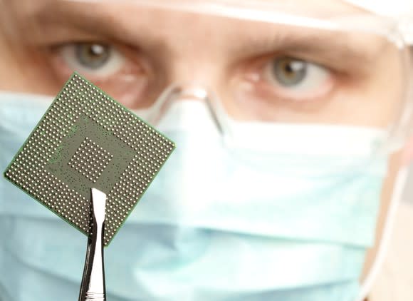 A technician in clean-room mask and goggles holds up a computer chip for a visual inspection.