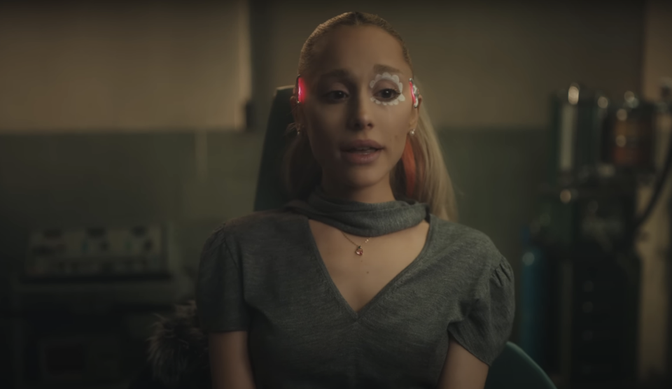 Ariana Grande with floral eye makeup and futuristic tech on her temples, wearing a turtleneck, in an exam chair