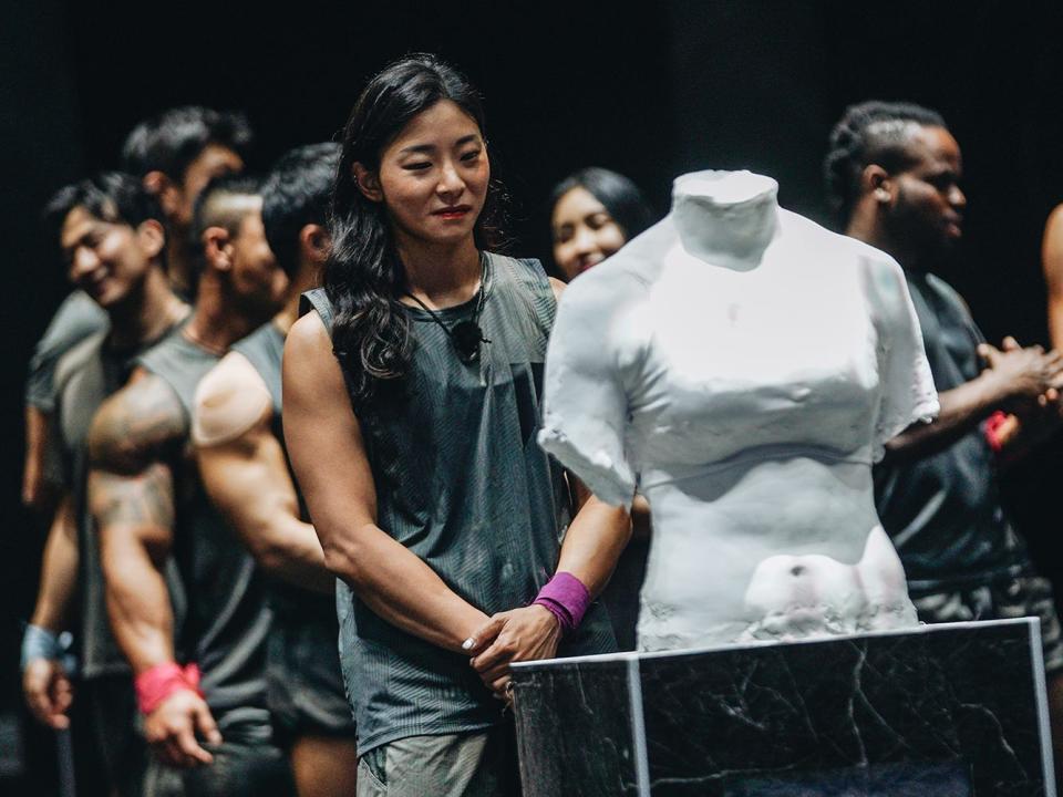 jang eun-sil on physical 100, standing in front of a plaster cast of her own torso with her hands folded in front of her. while the contestants standing behind her are chatting, jang appears reflective