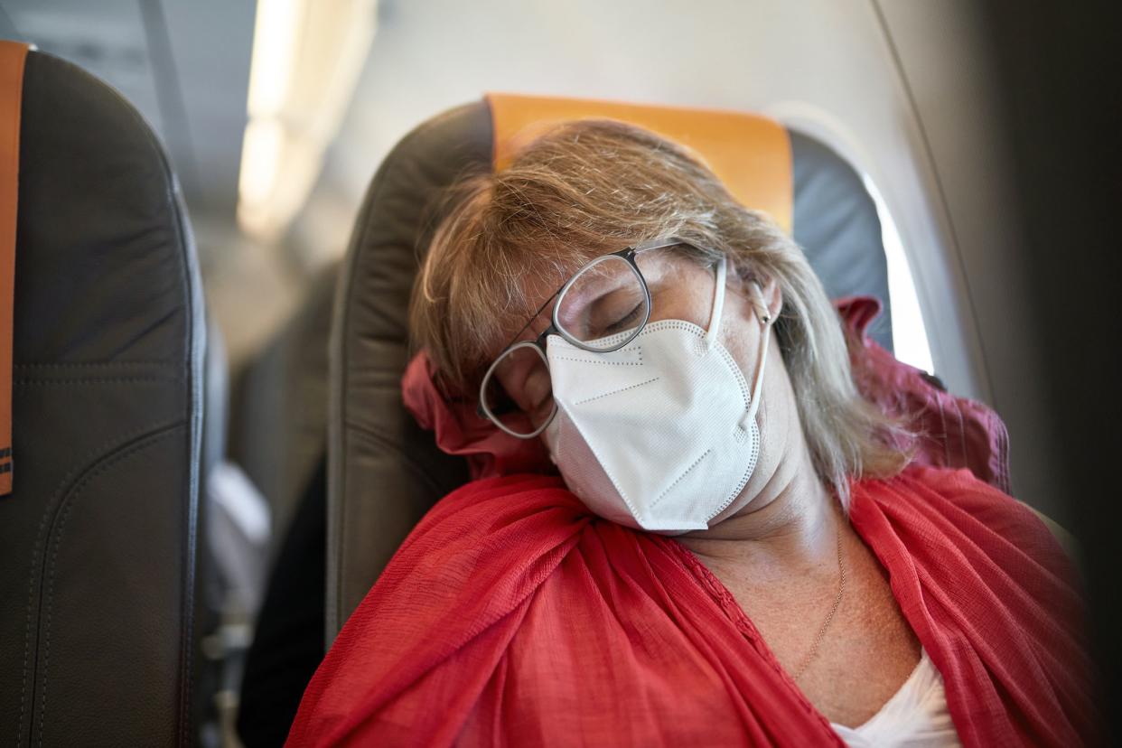 Front view of Caucasian woman in late 50s wearing eyeglasses, protective mask, and casual clothing asleep during flight in time of COVID-19.