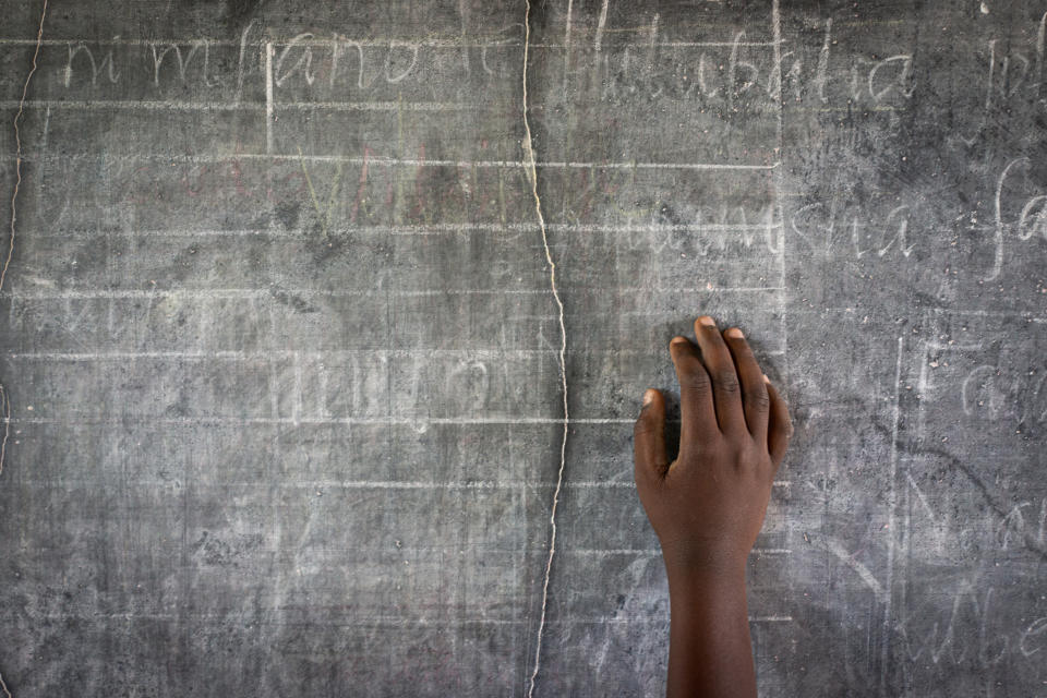 A child's hand is seen writing on a chalkboard with faint text