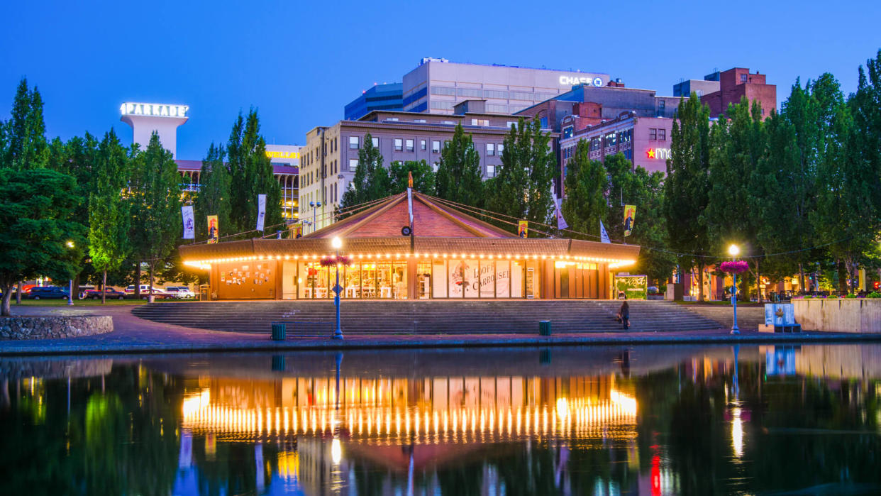 "Spokane, United States - July 21, 2012: People mill around outside the 1909 Looff Carousel, which is seen along the Spokane River with the downtown district of Spokane in the distance.