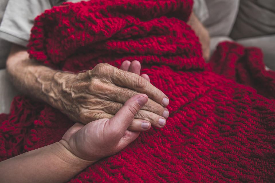 A younger person's hand gently holding an elderly person's hand over a red blanket