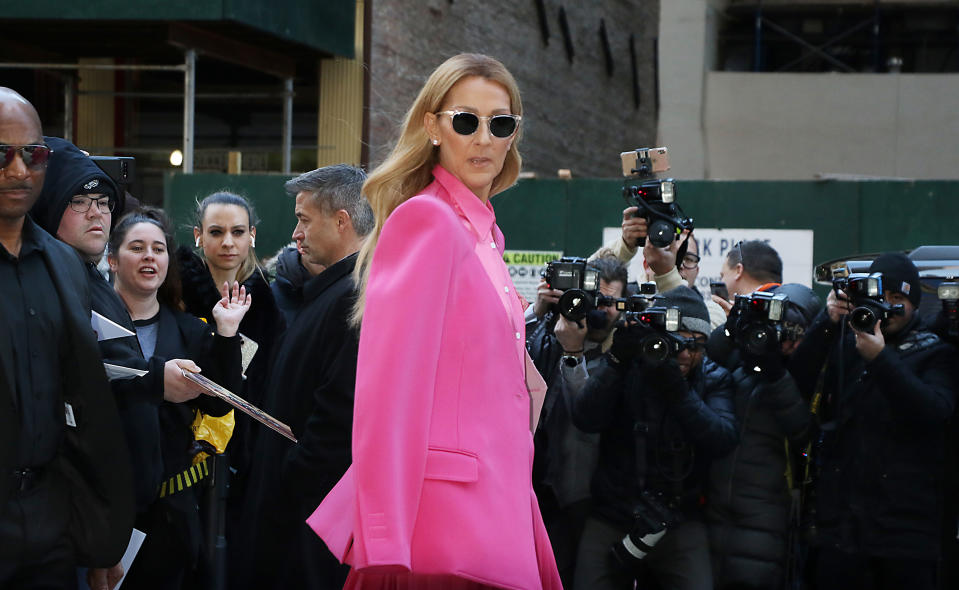 Celine Dion, wearing a stylish coat and sunglasses, is surrounded by photographers and fans