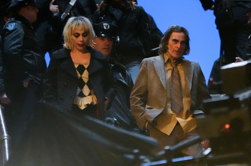 Lady Gaga and Joaquin Phoenix filming a scene surrounded by police officers. Lady Gaga wears a black and white checkered outfit, while Joaquin Phoenix wears a brown suit