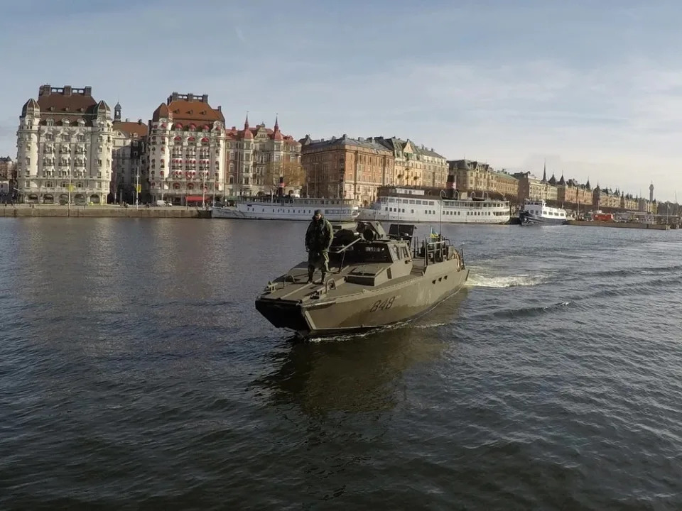 A Marine standing on the front of a CB90 as it sails near city buildings.