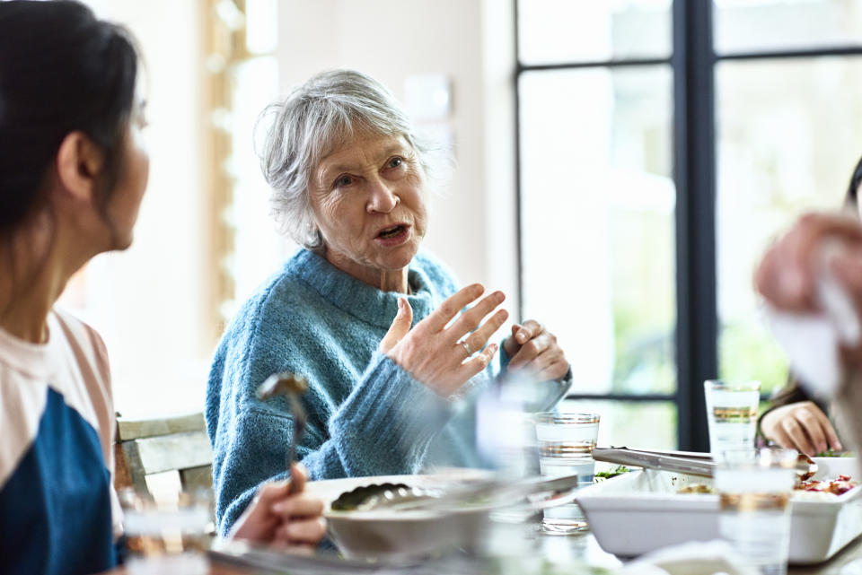 Candid portrait of woman in her 60s with grey hair, in conversation with friends, sitting at table with food and drink, woman listening in foreground