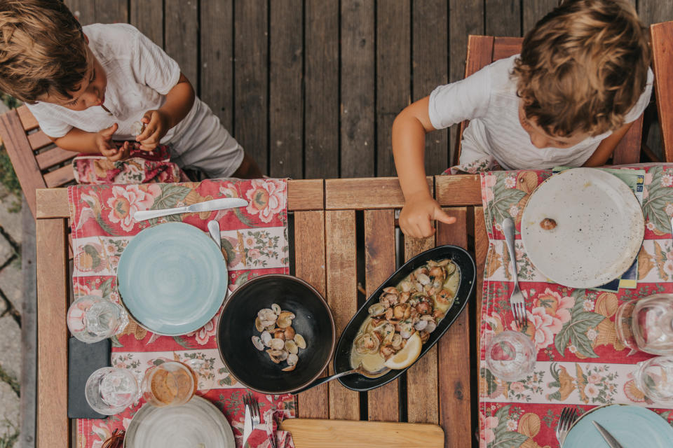 Two children at a wooden table with empty plates and a pan of cooked food