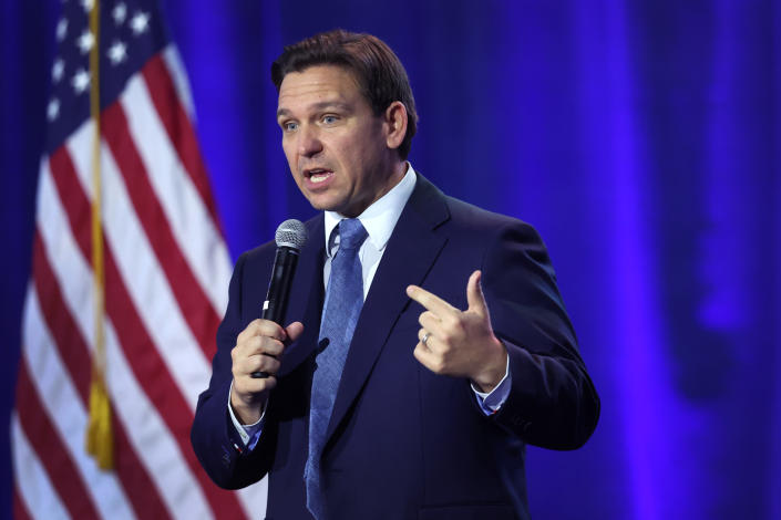 Florida Gov. Ron DeSantis holds a microphone to his mouth in front of a blue curtain and an American Flag.