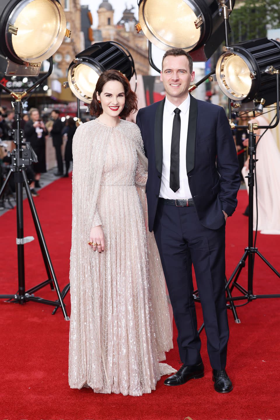 michelle dockery and jasper waller bridge stand smiling at the camera on the red carpet, she wears a long sequined gown and he a black suit