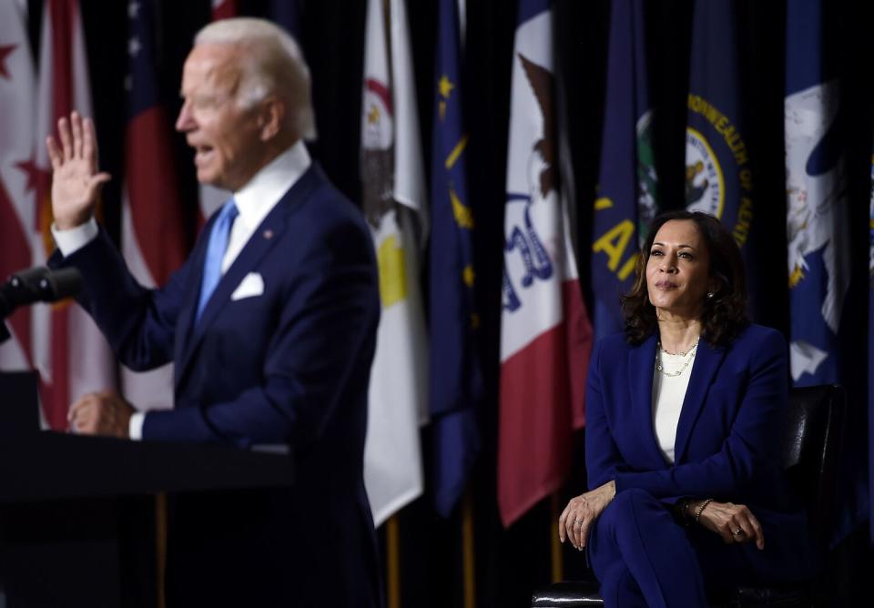 Joe Biden raising his hand and speaking from a lectern, as Kamala Harris sits behind him against a backdrop of various flags.