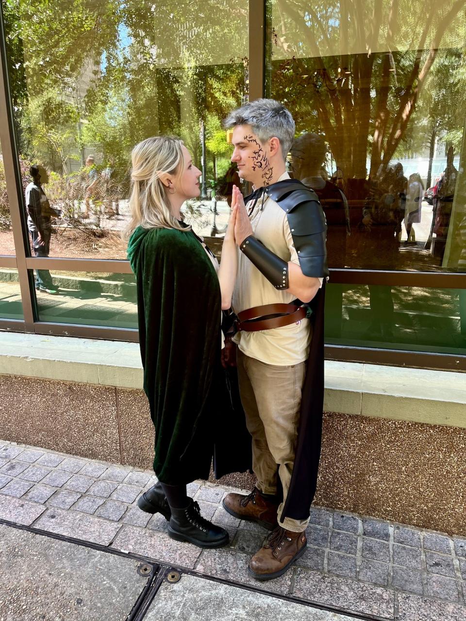 A woman in a green cloak presses her palm against a man's. He wears a cloak, has silver hair, and a face tattoo.