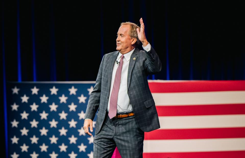 Texas Attorney General Ken Paxton, wearing a suit, waves in front of an oversized American flag