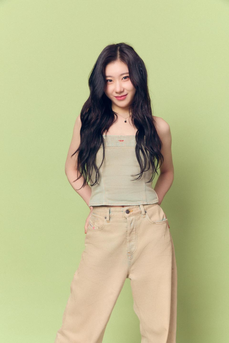 Chaeryeong from ITZY in a casual outfit with long, wavy hair, wearing a light tank top and pants, standing against a plain background