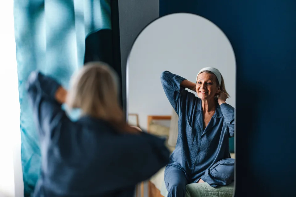Donna in pigiama sorride alla sua immagine riflessa nello specchio della camera da letto