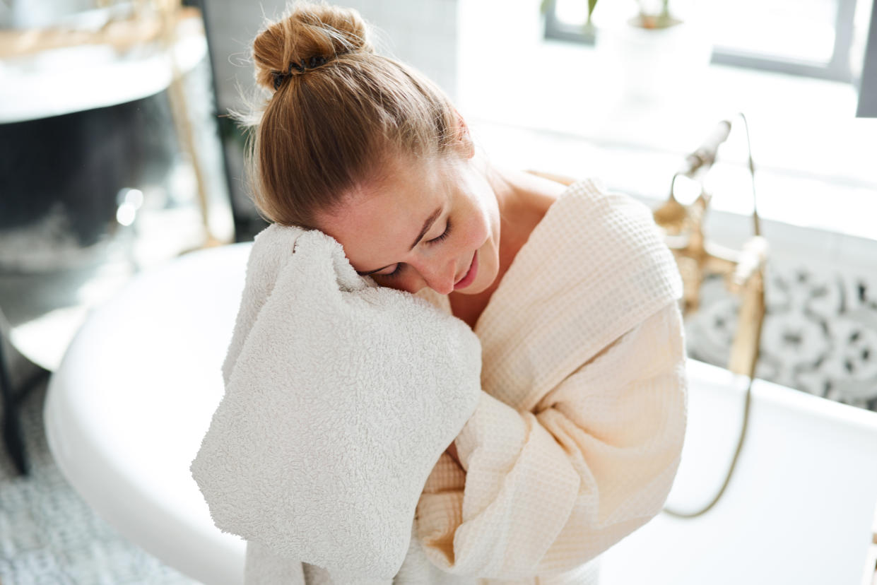                              A woman wearing a bathrobe presses her face into a white bath towel while standing in a bathroom