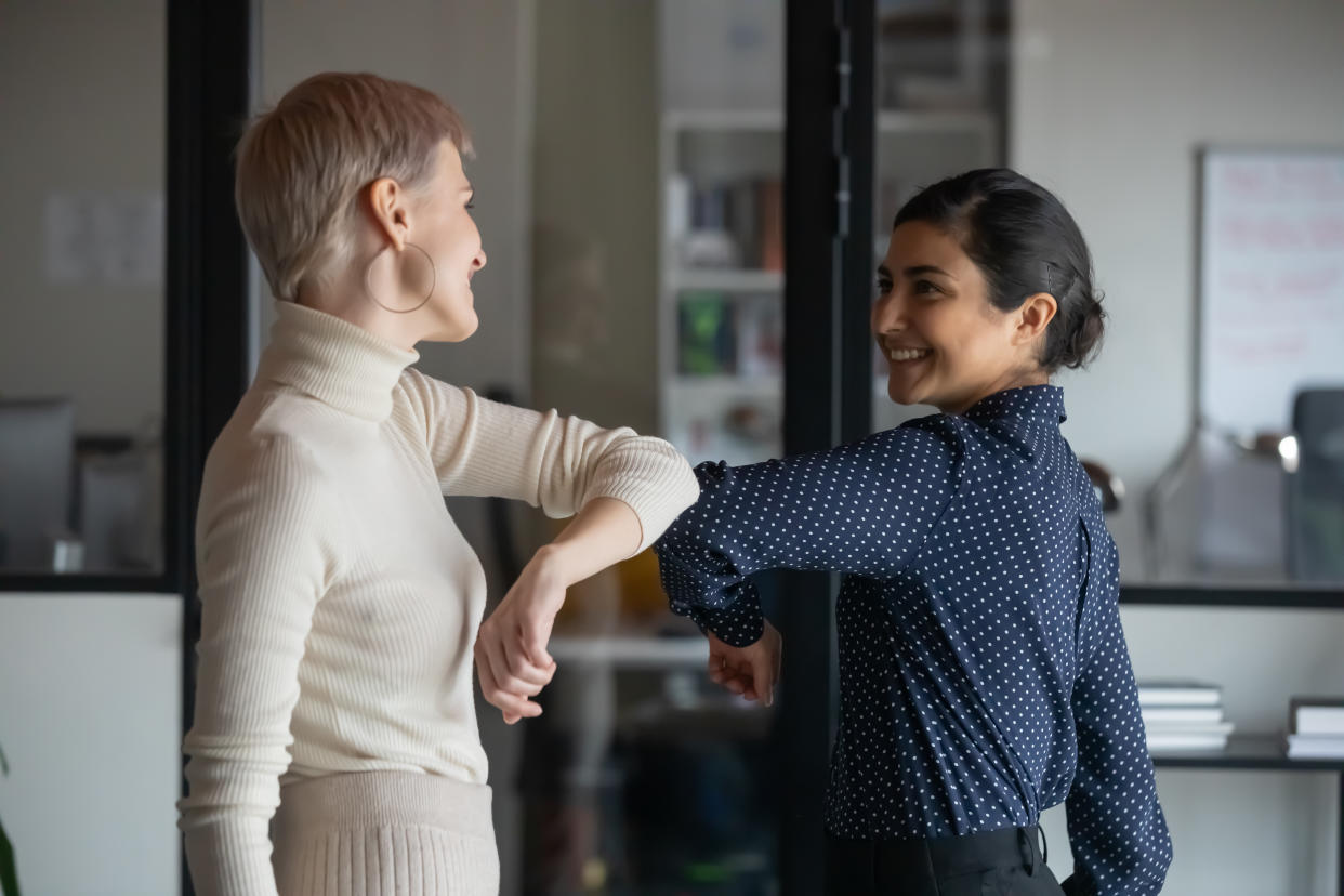 Friendly smiling millennial diverse female colleagues keeping social distance, greeting each other by bumping elbows instead of hugs or handshaking, preventing covid 19 coronavirus infection spread.