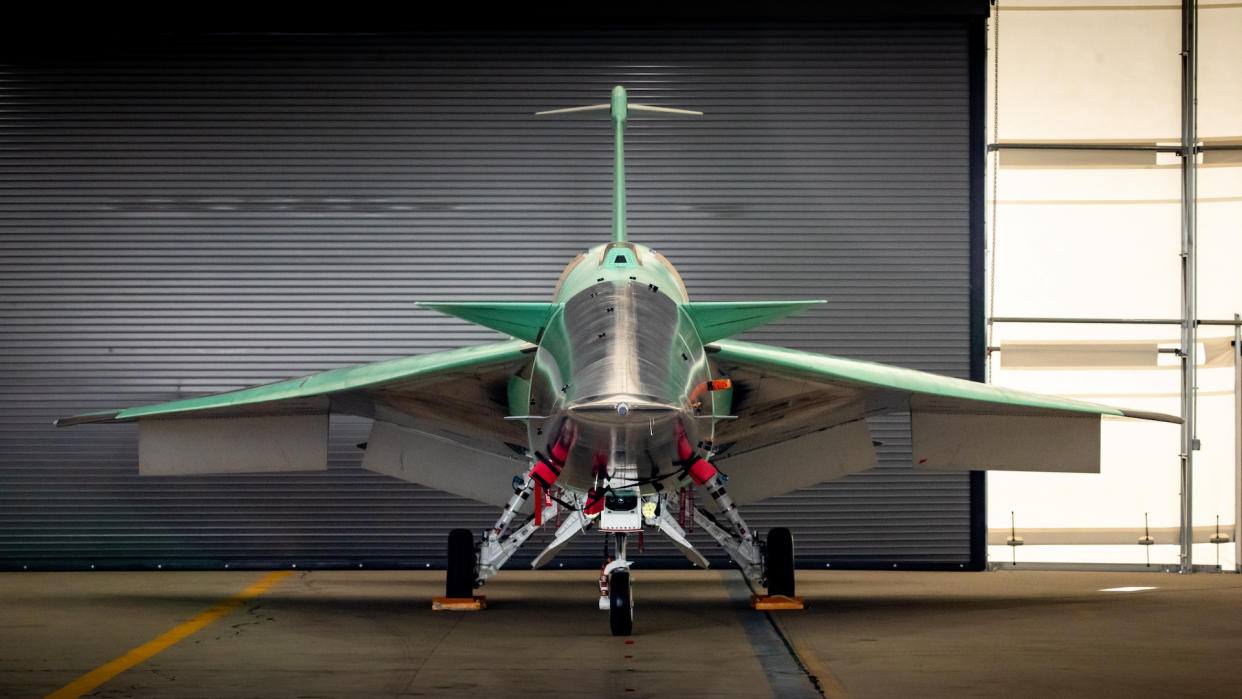  A head-on shot of NASA's X-59 supersonic jet, whose wingspan stretches across the middle of the photo. Green and black panels of the jet's body reveal sleek, sharp angles. The jet sits on three wheel connected by landing gear struts. The jet sits inside a hanger with white walls, and a grey corrugated door closed behind the jet. 