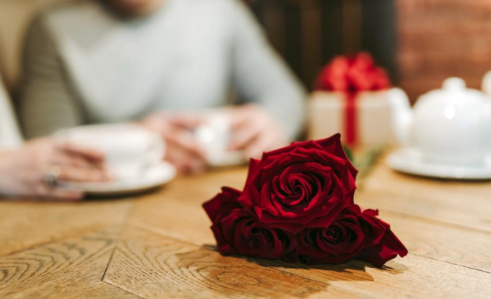 Two people are seated at a table on a date, blurred in the background, with teapots and cups; in focus, red roses are placed on the wooden table