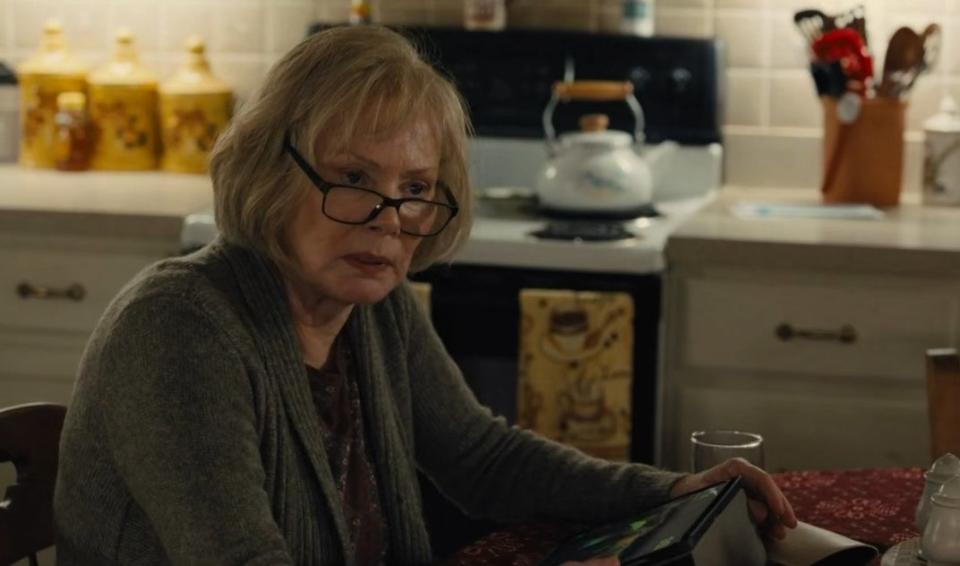 An elderly woman (Jean Smart) wearing glasses, sitting in a kitchen, and staring at something offscreen with a sullen look