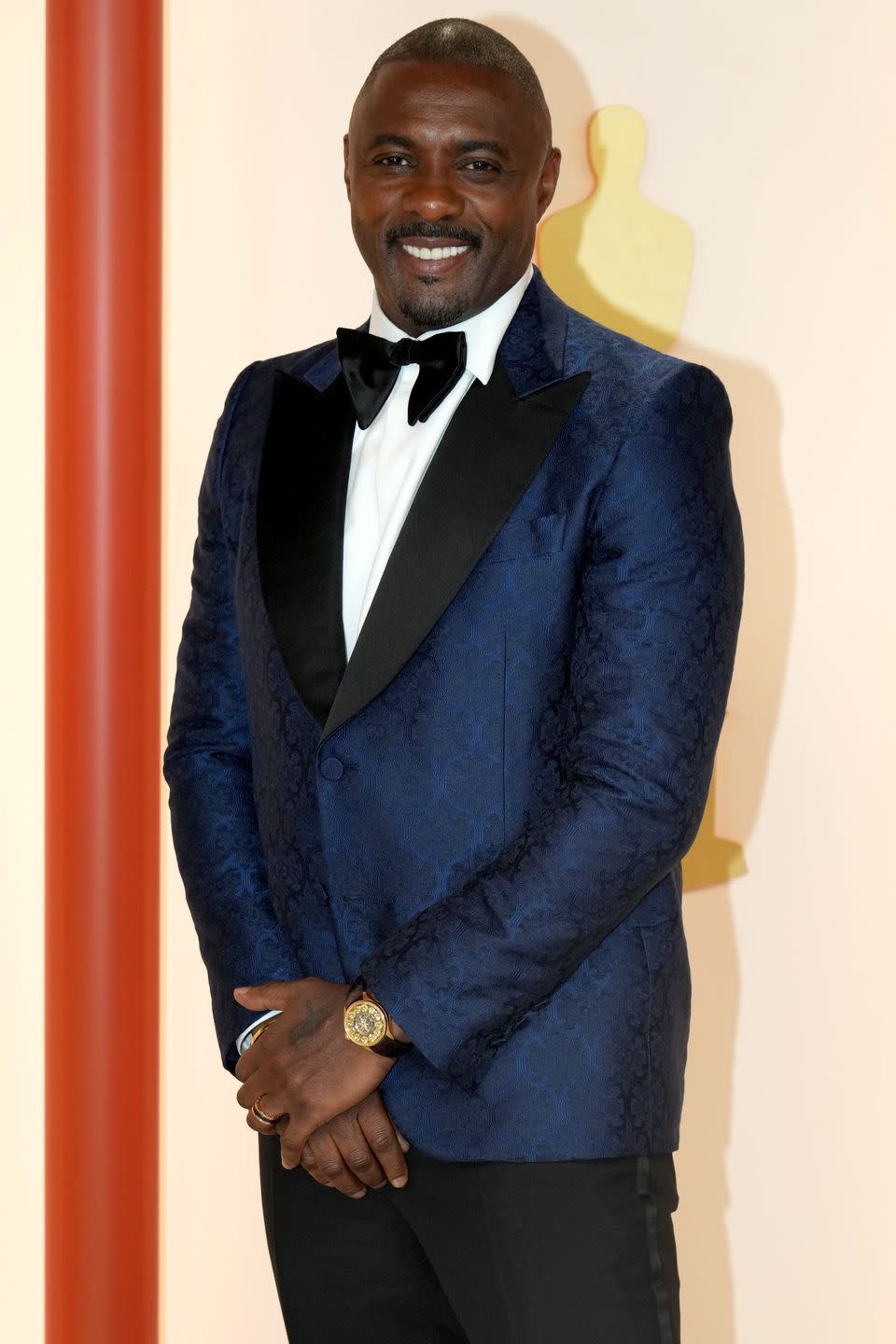 idris elba stands with hands clasped in front and smiling at the camera, closely shaved head with dark hair, wearing a blue tuxedo jacket with black trousers and bow tie and white shirt
