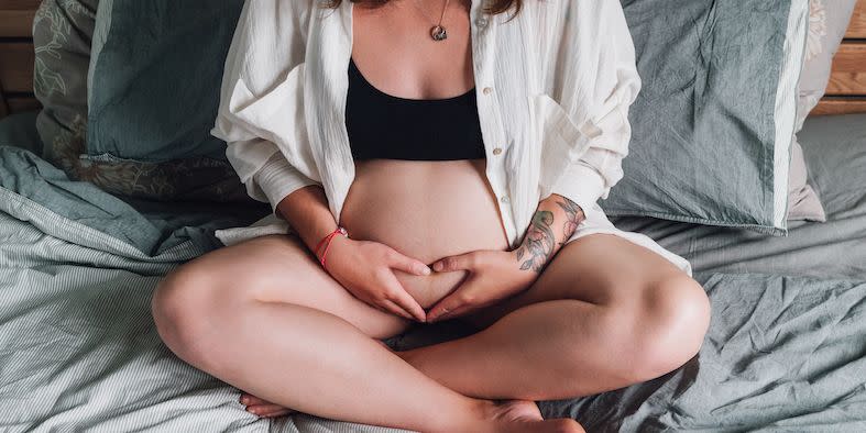 young pregnant woman with tattooed hand sitting cross legged touching her belly on a bed in soft light