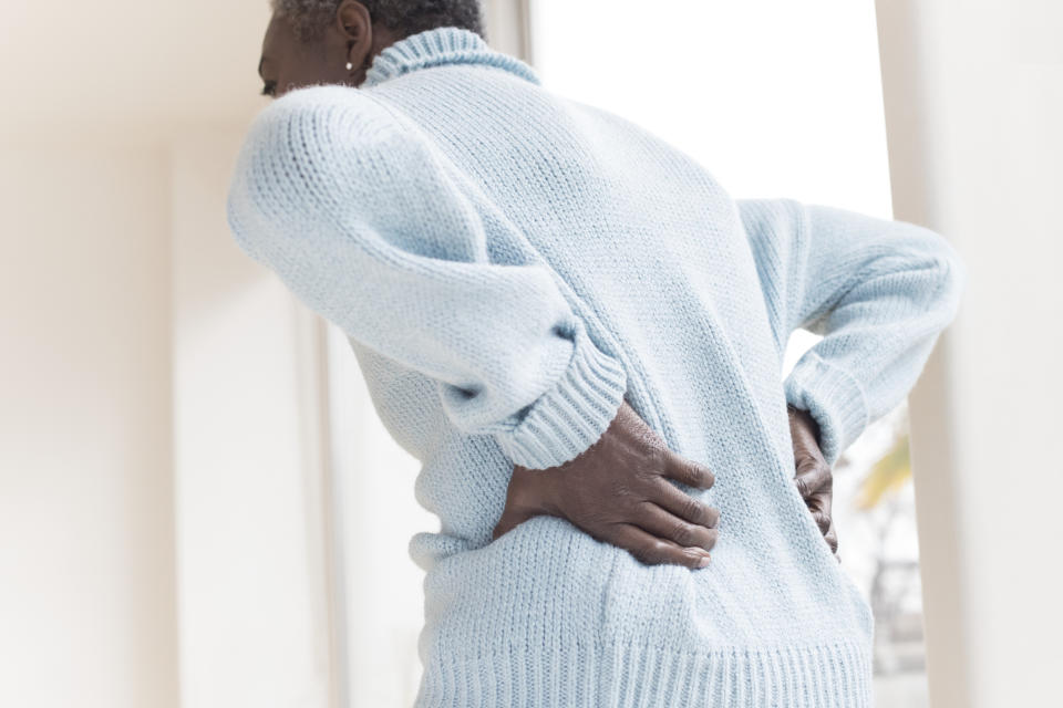 Person holding their lower back in pain while standing indoors, wearing a knitted sweater. The person in the image is not identified