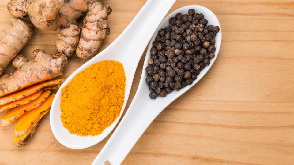 A spoon of ground turmeric beside a spoon of whole black pepper