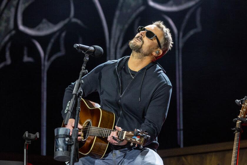 Eric Church, wearing sunglasses, looks up to the sky while sitting down on a stool on stage and playing an acoustic guitar