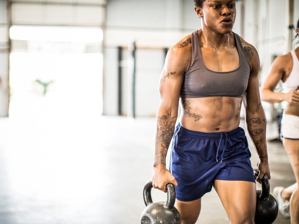 an athlete carrying two kettlebell weights in a farmer carry position in a gym