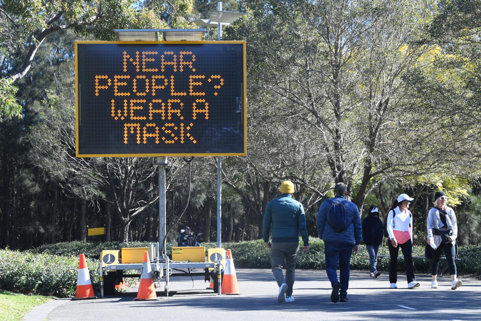 People walking on a path in a park with a digital sign reminding of the need to wear a mask.