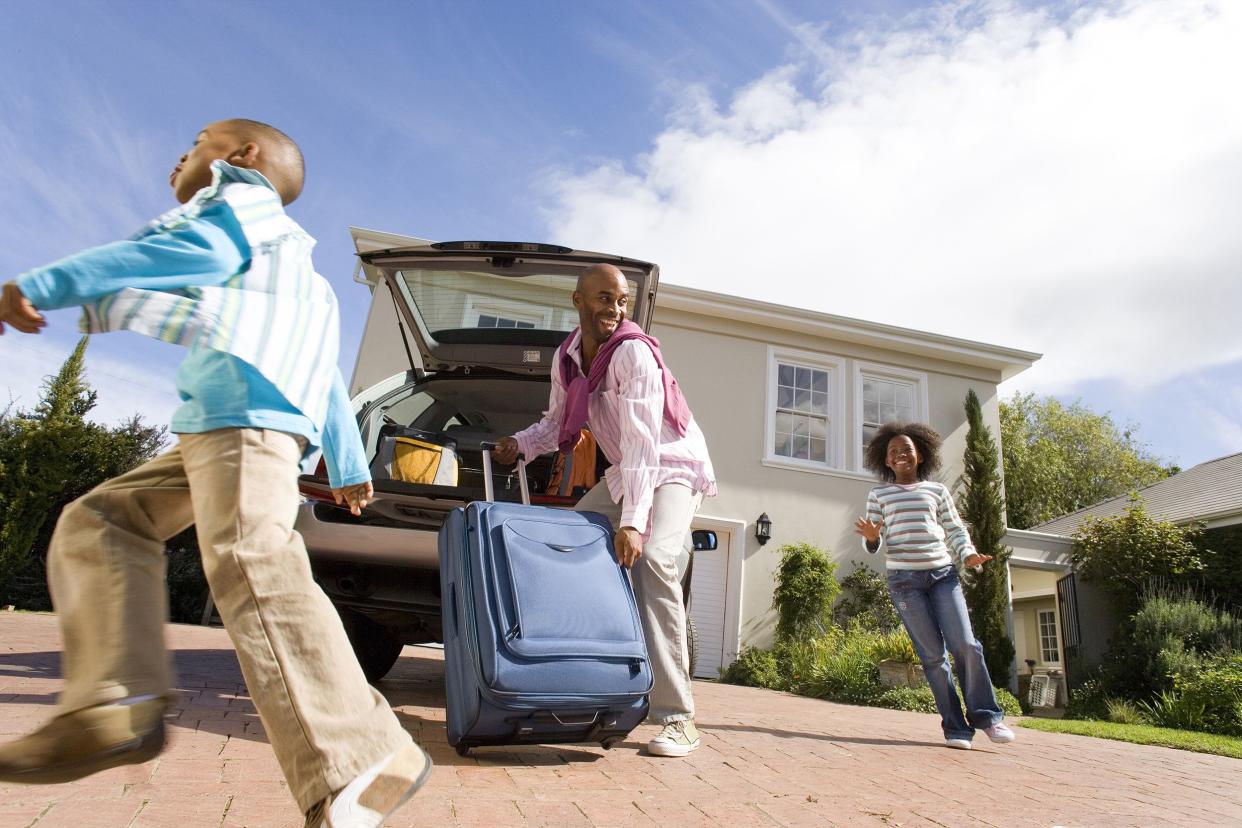 two children running by father packing suitcase into boot of car, smiling