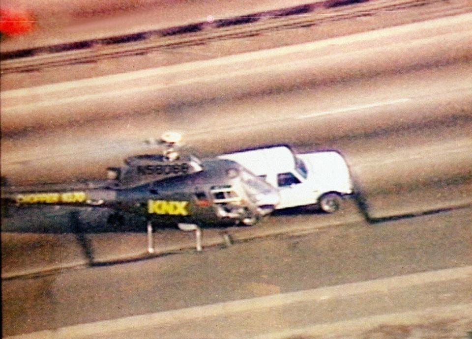 A helicopter flies above a white Ford Bronco along a highway.