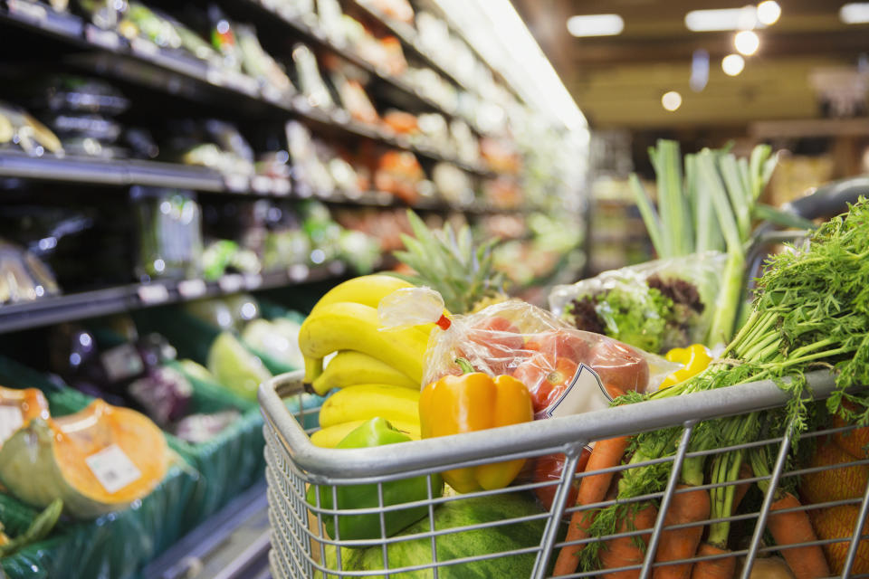 A grocery cart filled with fresh produce, including bananas, peppers, carrots, and other vegetables, in a supermarket aisle