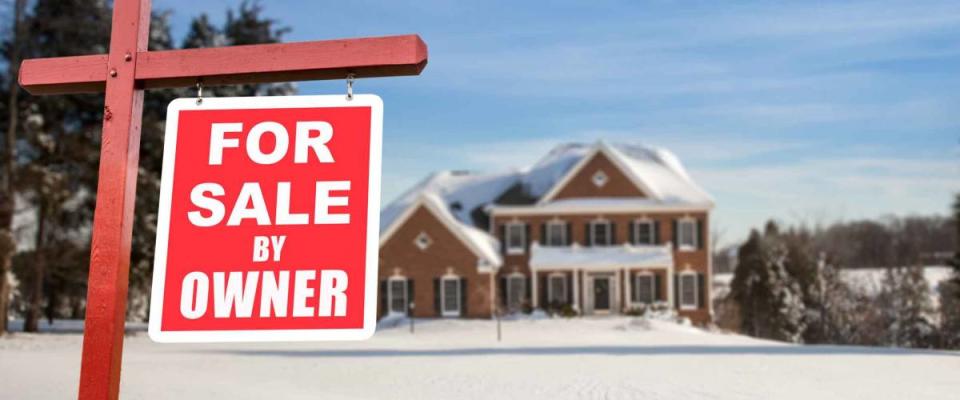 For Sale by owner real estate sign in front of large brick single family house in expansive snow covered yard in mid winter