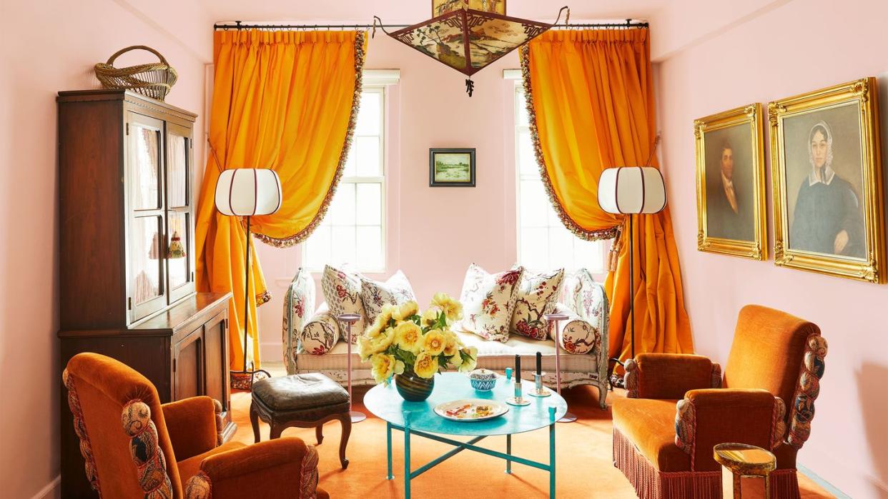 a bird and flower patterned daybed is behind saffron draped curtains and two floor lamps, in foreground is a turquoise cocktail table, two saffron velvet antique chairs, a cabinet, a pagoda pendant, and artworks