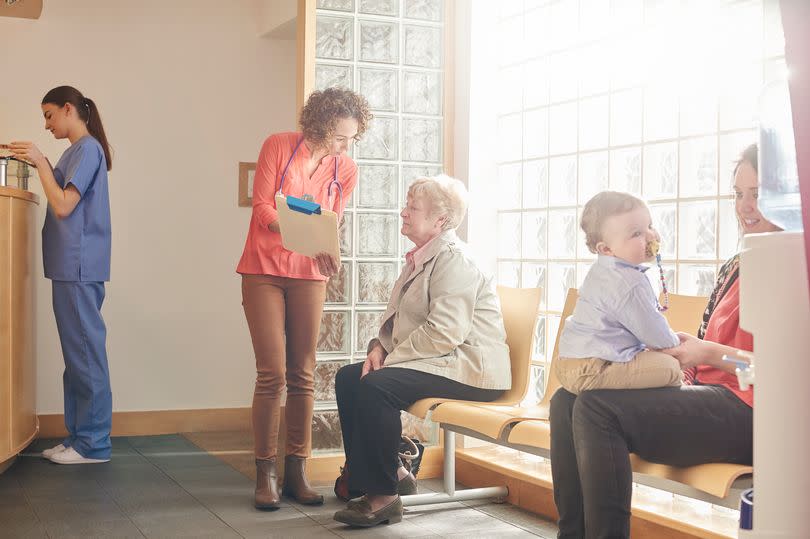 A female doctor stands next to a senior patient in her waiting room and casually chats through her factsheet . In the background a nurse stands at the reception desk whilst a mother and child play in the foreground waiting for their turn.